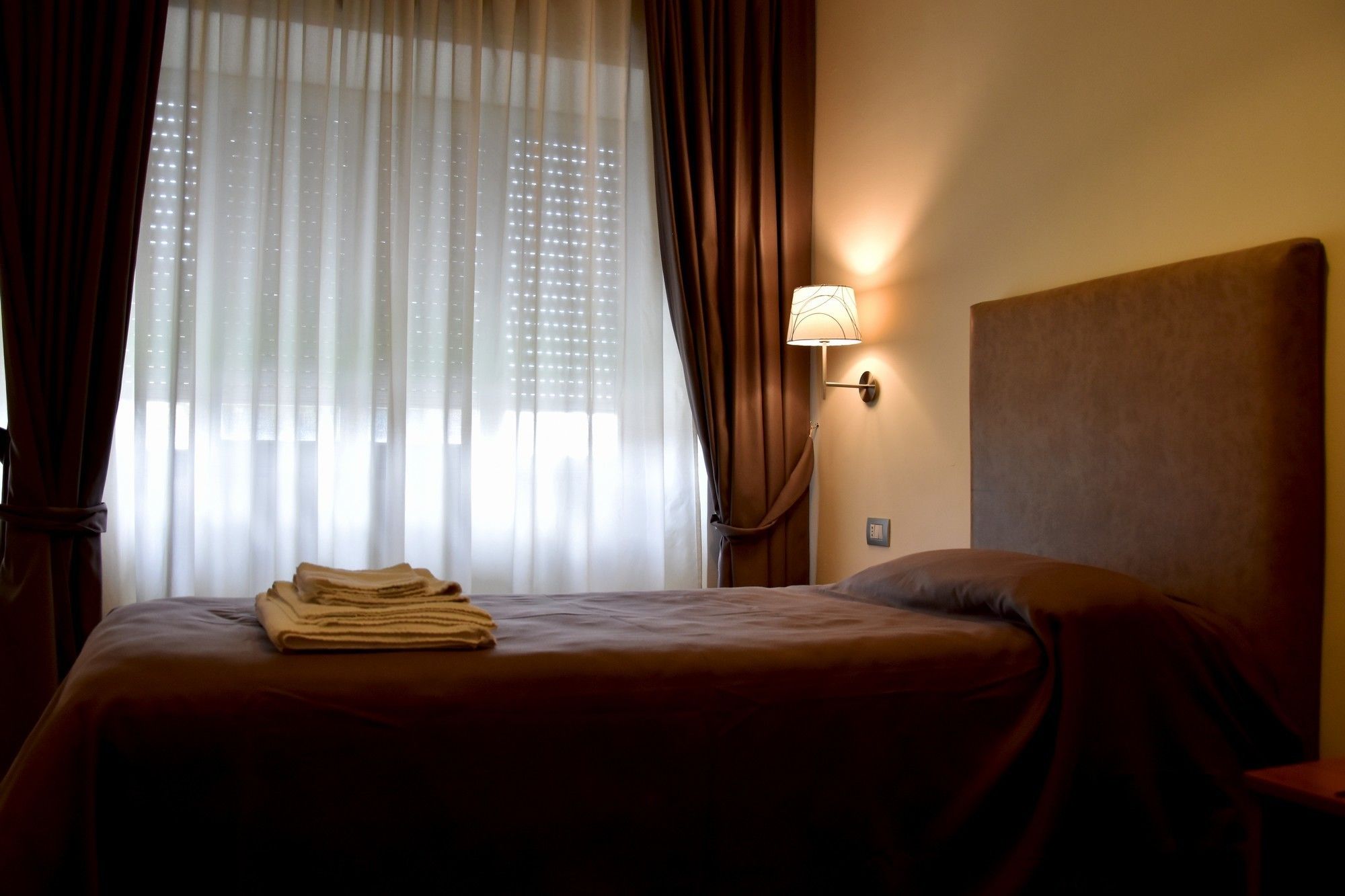 Une chambre d’hôtel avec un lit, une table de chevet, une lampe et des rideaux légers.
