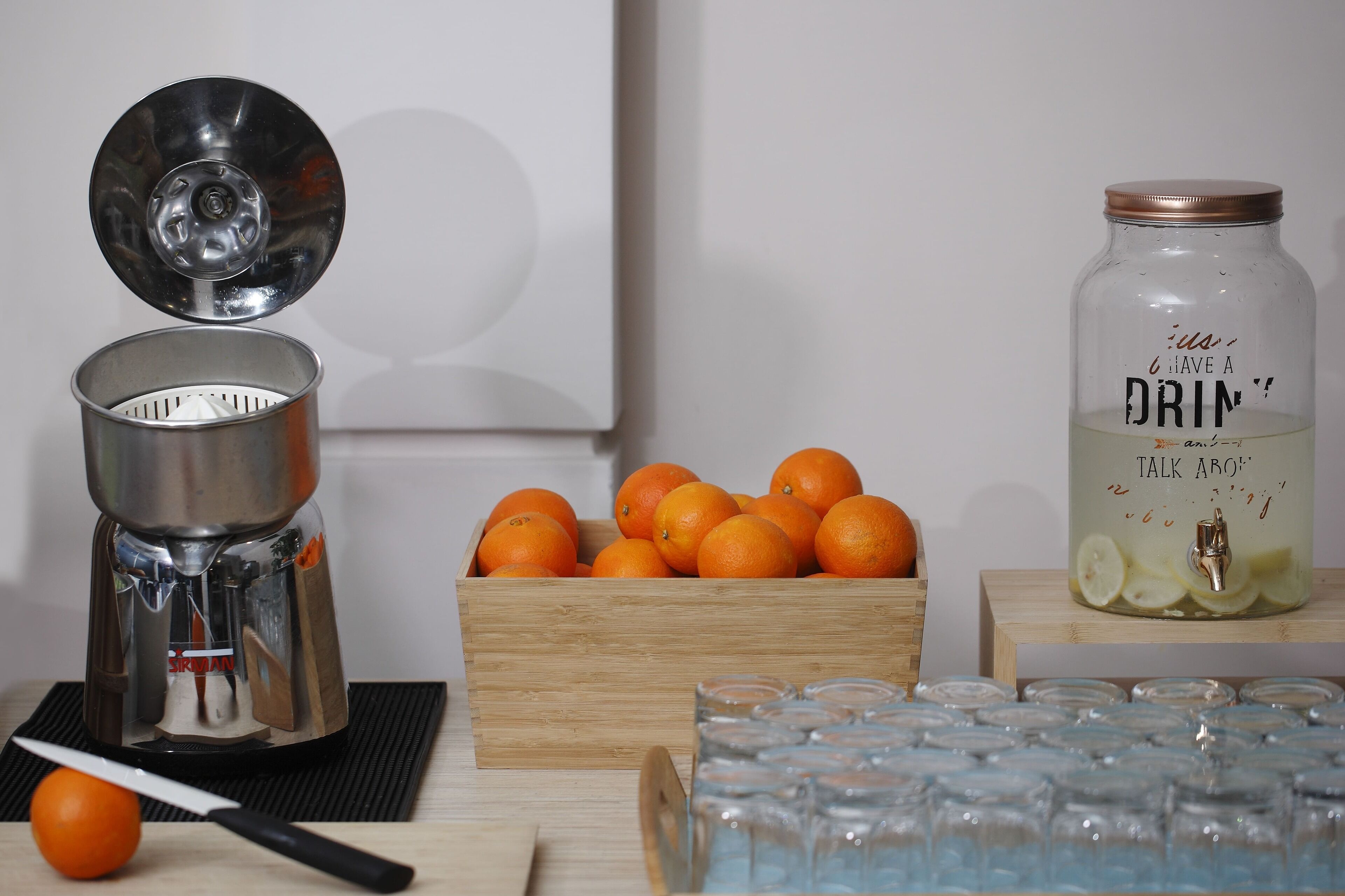 Een sapcentrifuge, een houten doos met sinaasappels, een waterdispenser en verschillende glazen op een tafel.