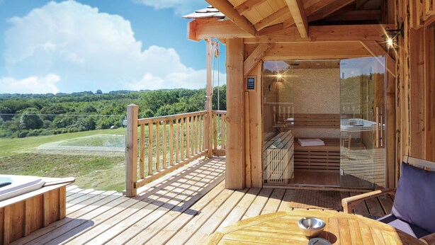 Een houten terras met een jacuzzi, een glazen sauna en uitzicht op een groen landschap.