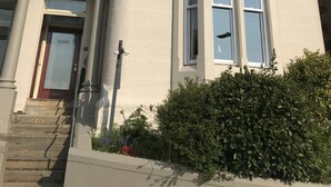 Un edificio de dos pisos con fachada color crema, una puerta roja y un pequeño jardín con arbustos y flores.