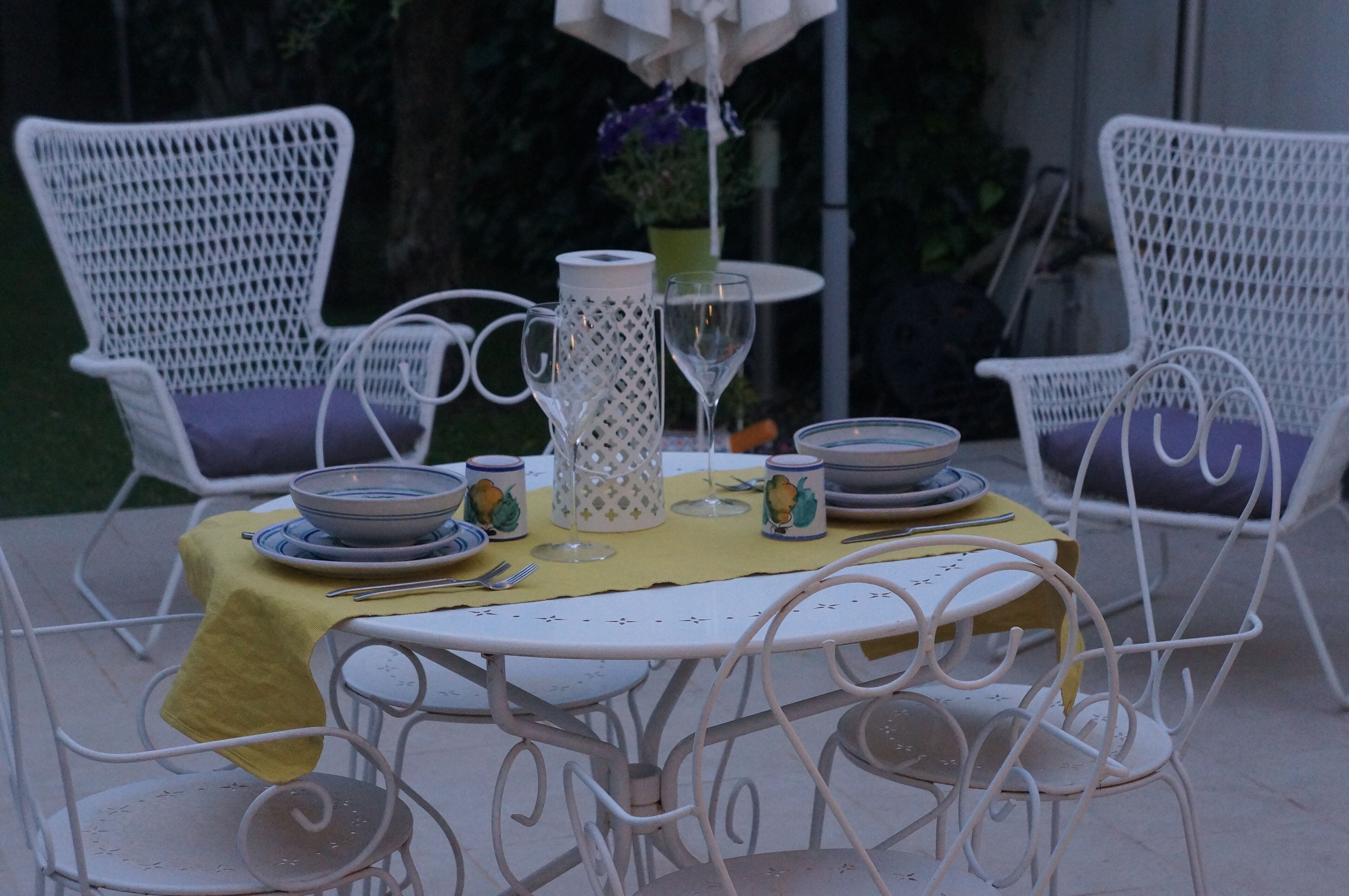 Een tafel buiten gedekt, met witte rotanstoelen, een gele tafelkleed en een witte kruik.