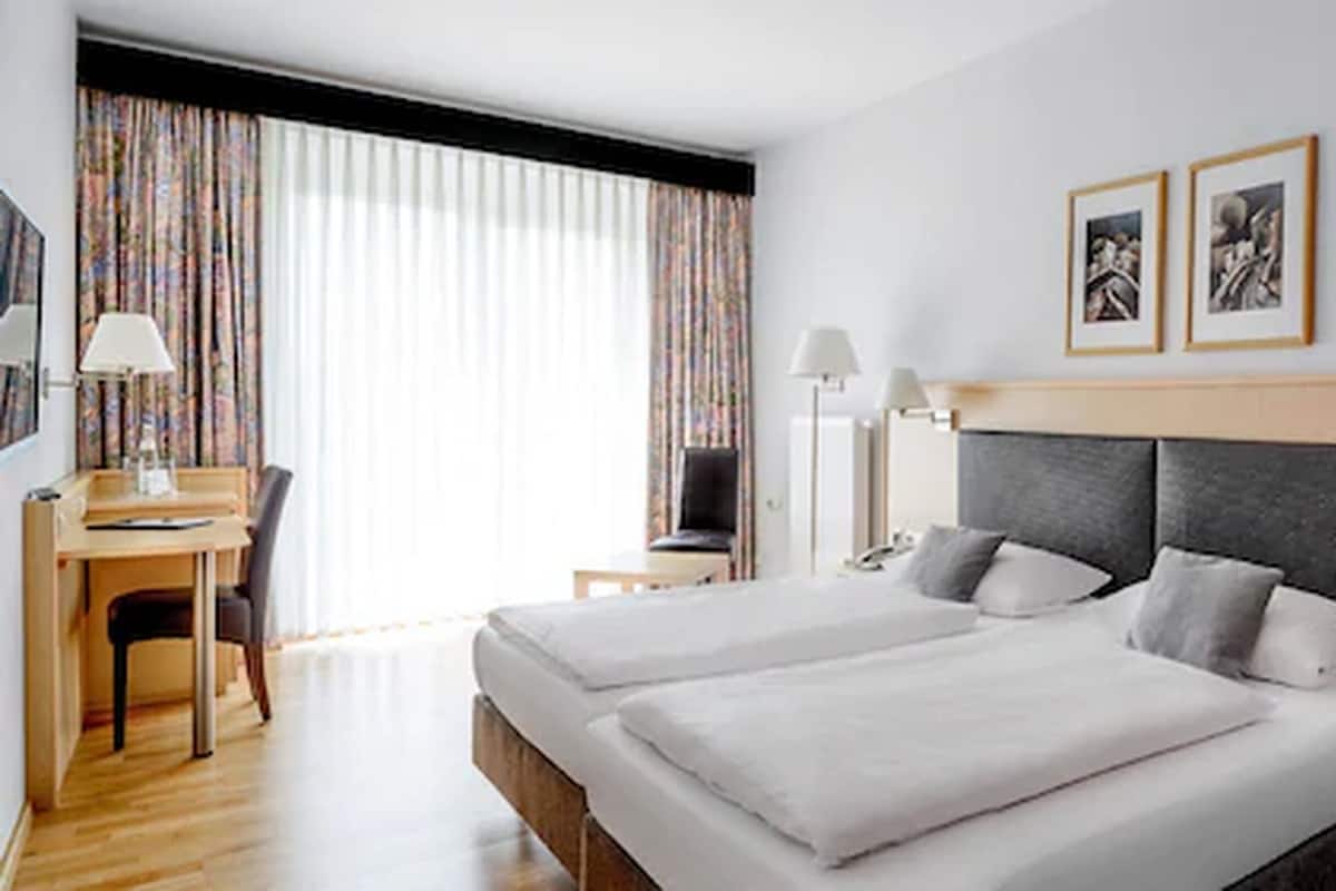 A hotel room with a double bed, a desk with a chair, a lamp, and two framed pictures on the wall.