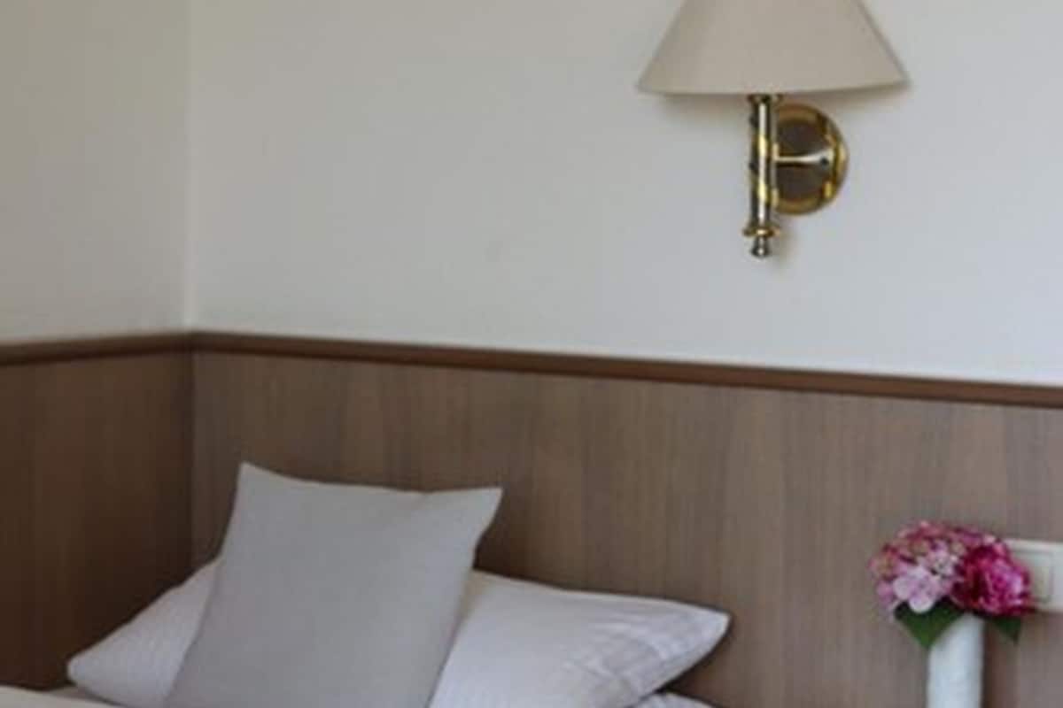 A neatly made bed with white linens, a wooden headboard, a bedside table with a vase of flowers, and a wall-mounted lamp.