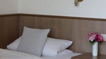 Una cama bien hecha con sábanas blancas, cabecera de madera, mesita de noche con un florero y una lámpara fijada a la pared.