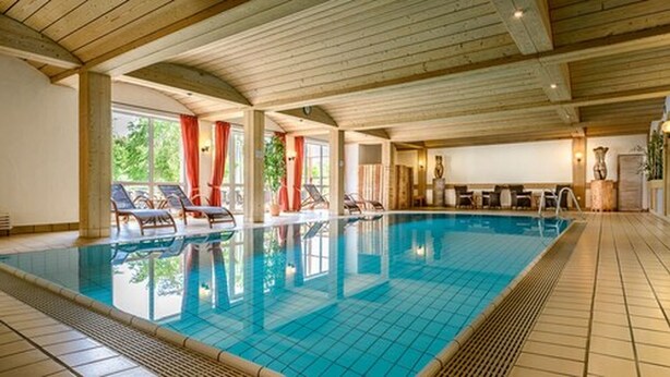 Una piscina interior con paredes y techo de azulejos, rodeada de sillones de descanso y suelo de madera.