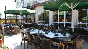 Un espacio exterior para comer con mesas y sillas, sombrillas verdes y un edificio con grandes ventanales al fondo.