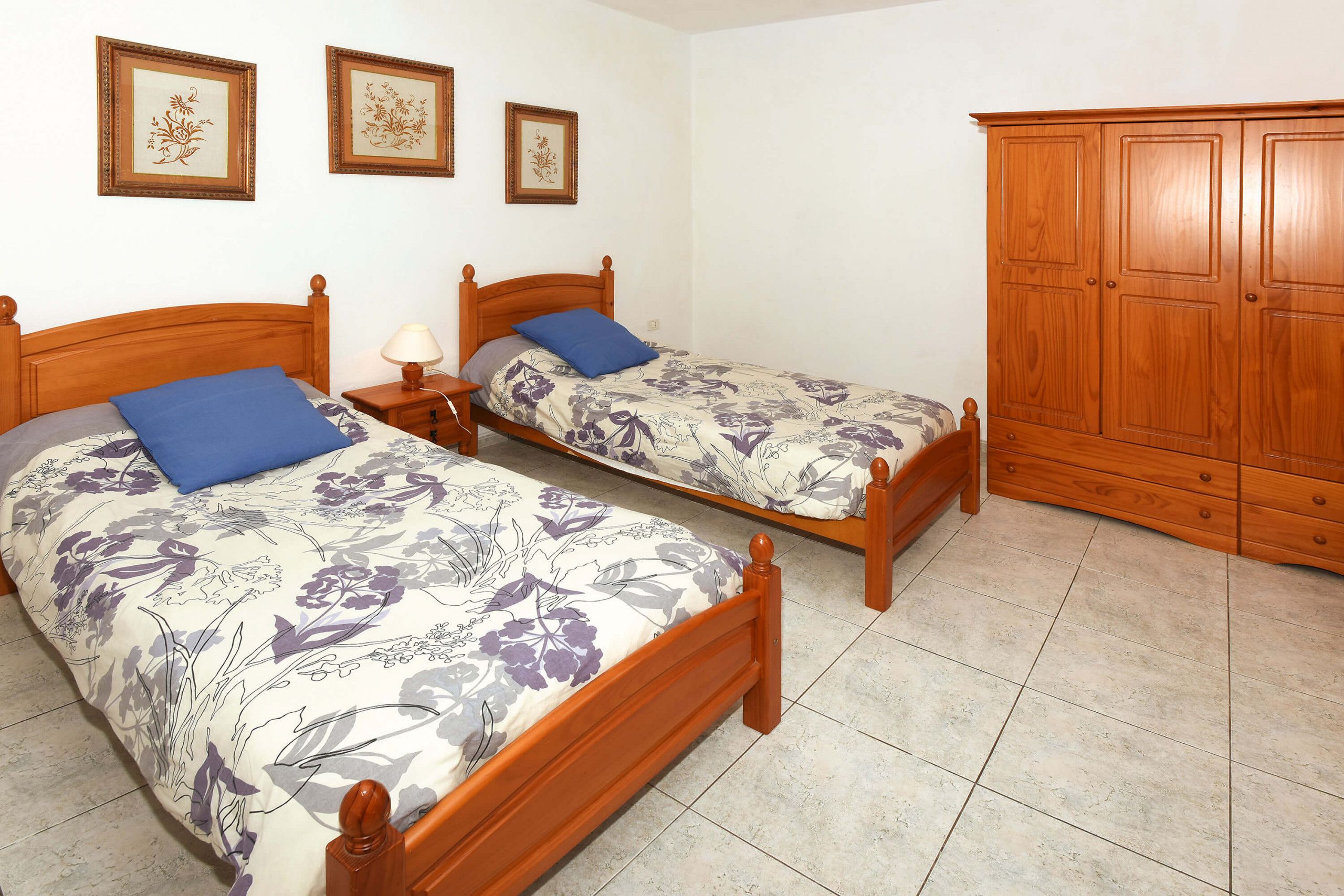 Two wooden beds with floral bedding, a wooden wardrobe, and a tiled floor.
