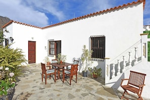 A white building with a tiled roof, a wooden table with chairs, and potted plants.