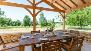 Una pérgola de madera al aire libre con una mesa de comedor dispuesta para cuatro personas, rodeada de muros de piedra y vistas a un paisaje cubierto de césped.