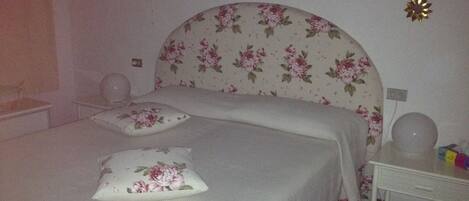 Un dormitorio con cabecera floral, dos almohadas, una mesita de noche con lámpara y un pequeño atril.