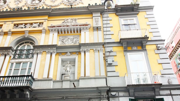 A historic building with a yellow facade, ornate decorations, and a black door with a sign. - Le Quinte del Bellini (Naples)