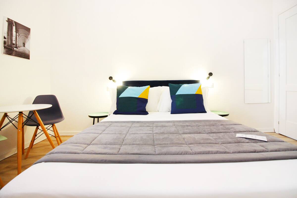 A neatly made bed with a grey duvet, two pillows with geometric patterns, a small round table, and a chair with wooden legs.