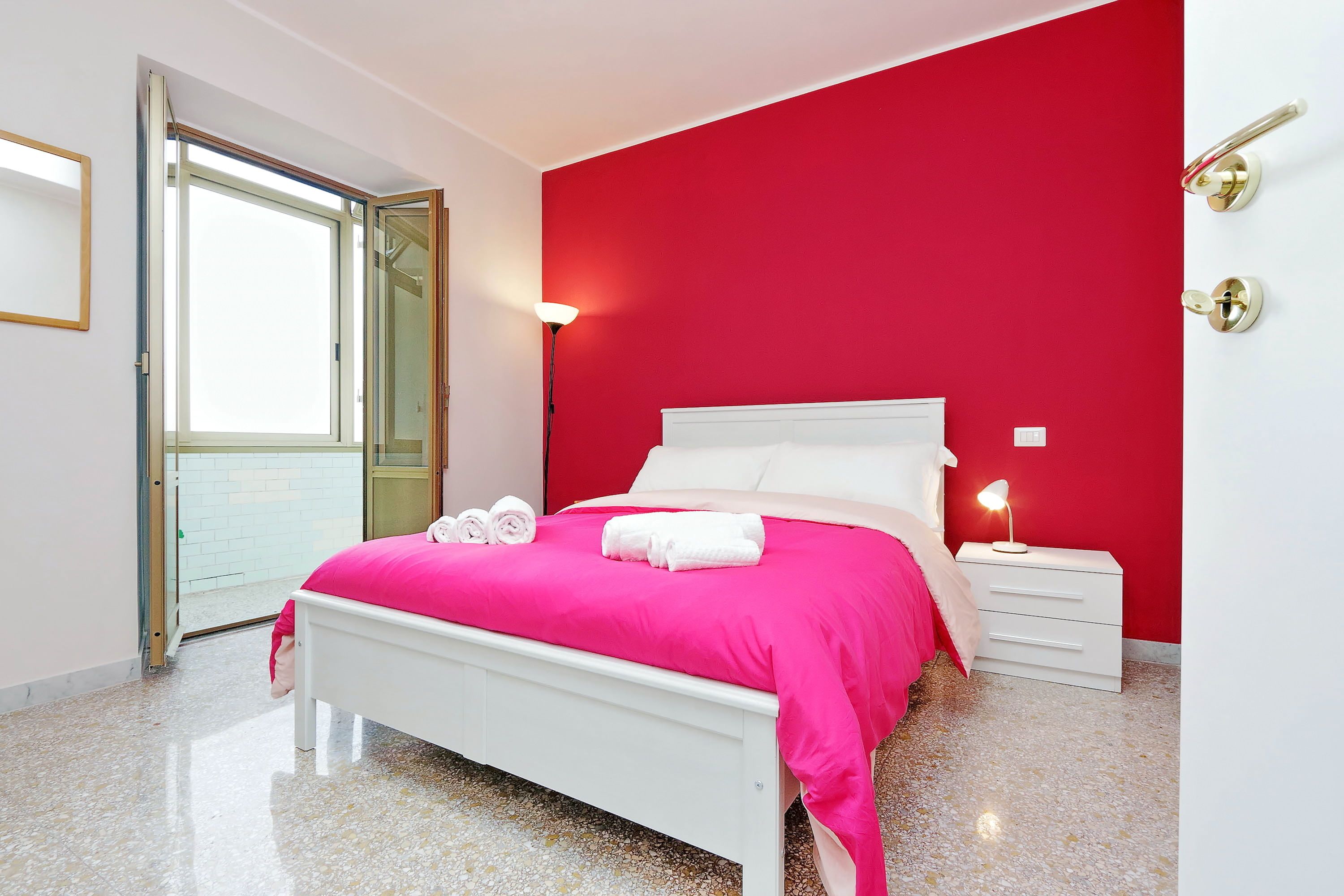A bedroom with a white bed, red walls, a white nightstand, and a door leading to an outdoor area.