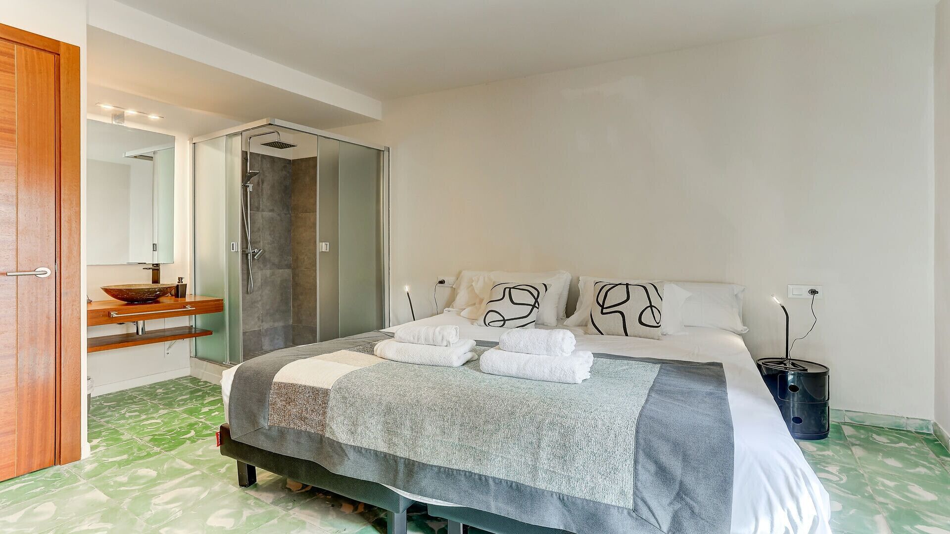 Una habitación de hotel moderna con una cama grande, un baño con ducha, un tocador de madera y piso de baldosas verdes.