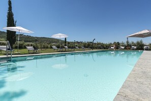 A clear swimming pool with lounge chairs and umbrellas, surrounded by greenery and hills.