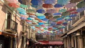 A bustling street market with colorful umbrellas overhead, wooden tables and chairs, and vendors selling goods.