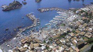 An aerial view of a coastal town with a marina, rocky outcrops, and a dense urban area with buildings and parked cars.