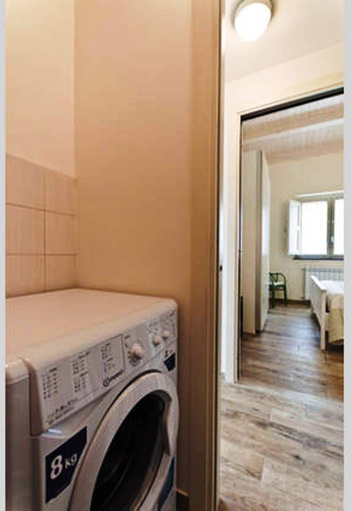 A laundry room with a front-loading washing machine, wooden flooring, and a view into a bedroom with a bed and a window.