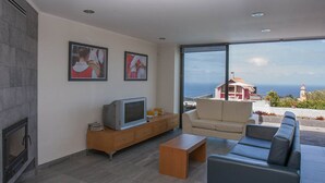 A modern living room with a fireplace, a television, and a sofa. There are two framed pictures on the wall and a view of the sea and a building outside. - A Casa do Lagar In a peaceful and solitary area with a pleasant climate all year (São Jorge)