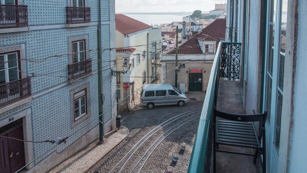 Een balkon met uitzicht op een geplaveid straatje, een tramrails, gebouwen met balkons en een verre zeespiegel.