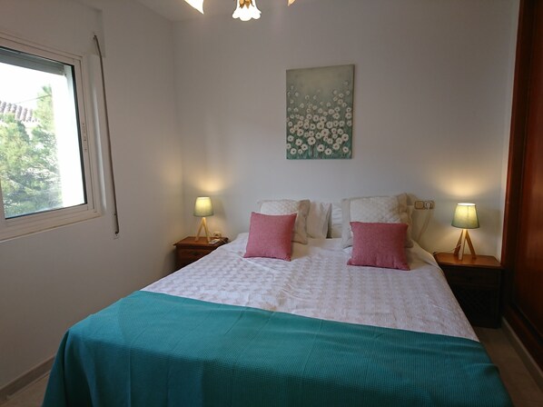 A bedroom with a bed, two bedside tables with lamps, a window, and a floral painting on the wall. - Near the Sea, Views and Golf Next to the Sea and Golf at the Lighthouse. (El Faro)