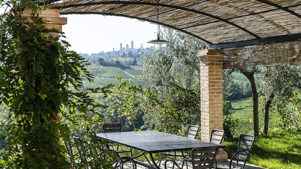 An outdoor patio with a pergola, a table, and chairs, overlooking a vineyard and a distant city skyline. - Martina, apartment with terrace on the green hills of San Giminiano (San Gimignano)