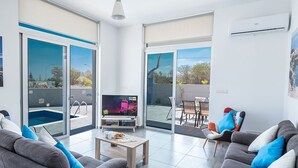 A modern living room with a TV, sofa, coffee table, and a view of a swimming pool through large glass doors. - Villa Desi, Luxury and new 5BDR Protaras Villa with pool and stunning sea views (Protaras)