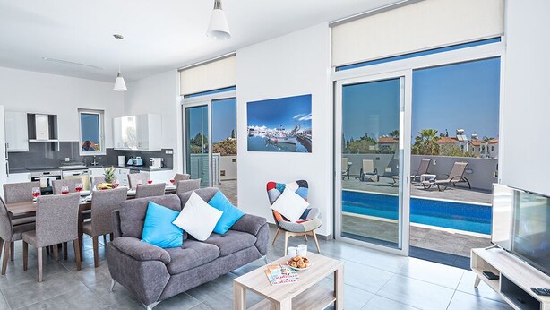 A modern living room with a grey sofa, a wooden coffee table, and a TV. There is a dining area with chairs and a table, and a kitchen in the background. - Villa Desi, Luxury and new 5BDR Protaras Villa with pool and stunning sea views (Protaras)
