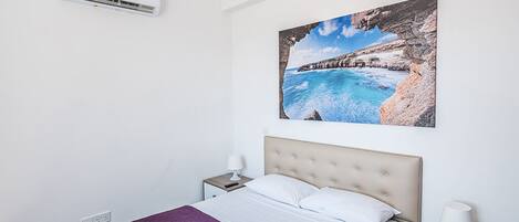 A hotel room with a bed, a painting of a coastal landscape, an air conditioning unit, and a nightstand with a lamp.