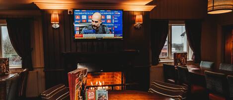 A cozy bar area with a wooden table, striped chairs, a television displaying a sports event, and a fireplace.