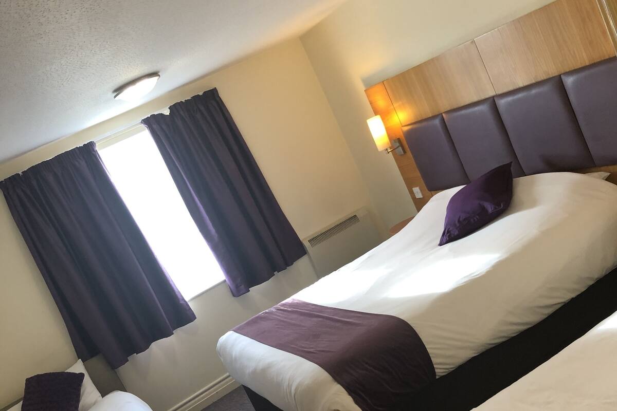 A hotel room with a large bed, a smaller bed, and a desk. There is a window with curtains, a headboard, and a nightstand with a lamp.