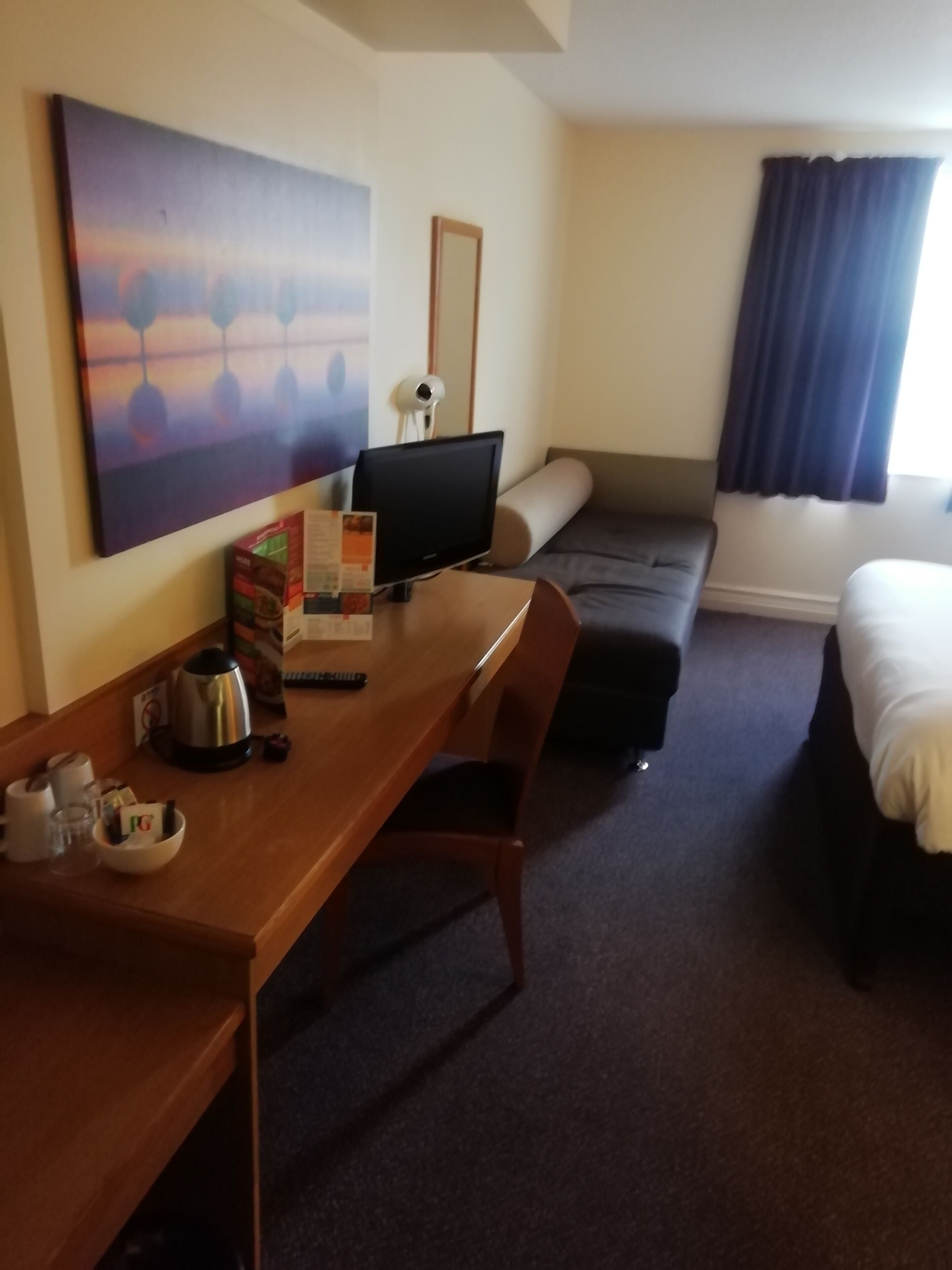 A hotel room with a wooden desk, a television, a sofa, a bed, and a painting on the wall.