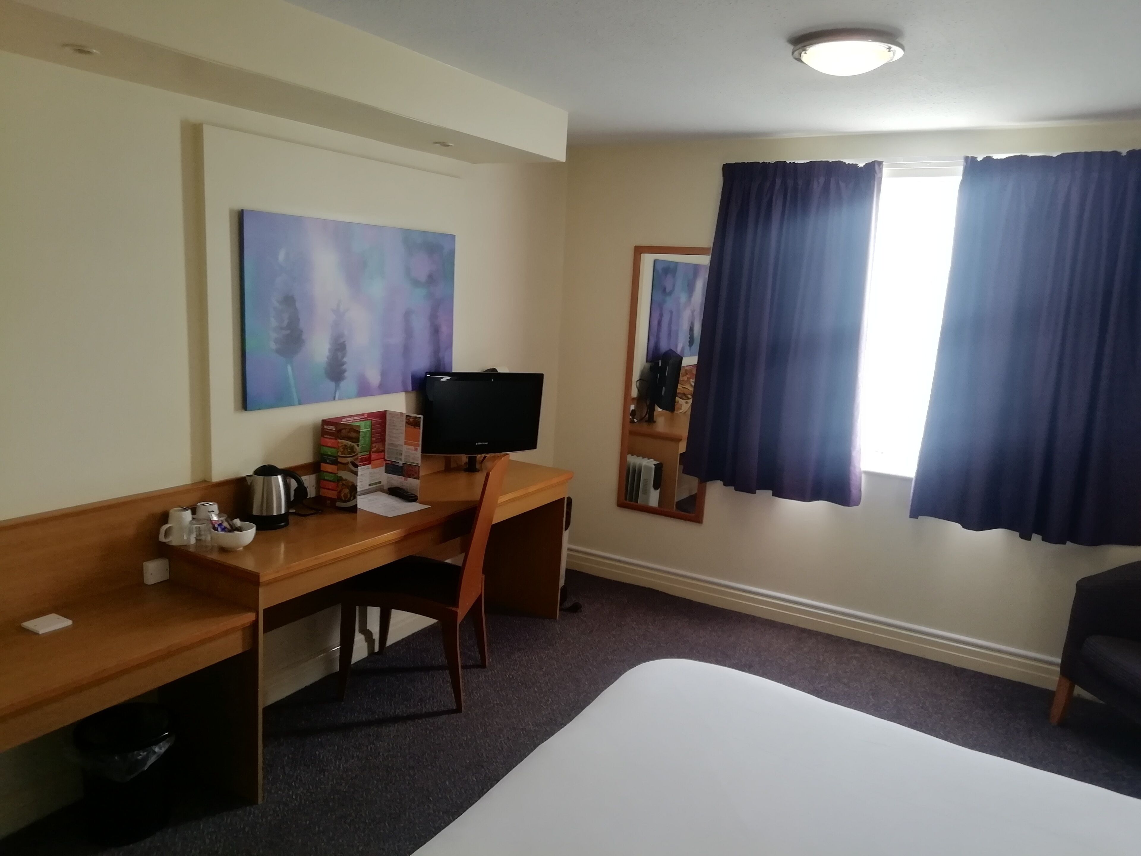 Une chambre d’hôtel avec un bureau en bois, un ordinateur, un lit, une fenêtre avec des rideaux et un tableau au mur.