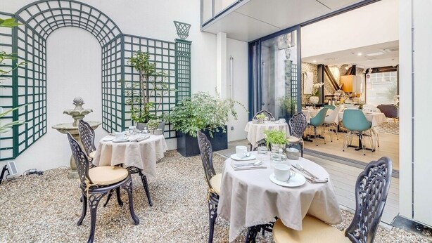 An outdoor dining area with tables and chairs, surrounded by decorative plants and a stone fountain.