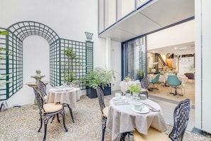 Un espacio de comedor al aire libre con mesas y sillas, rodeado de plantas decorativas y una fuente de piedra.