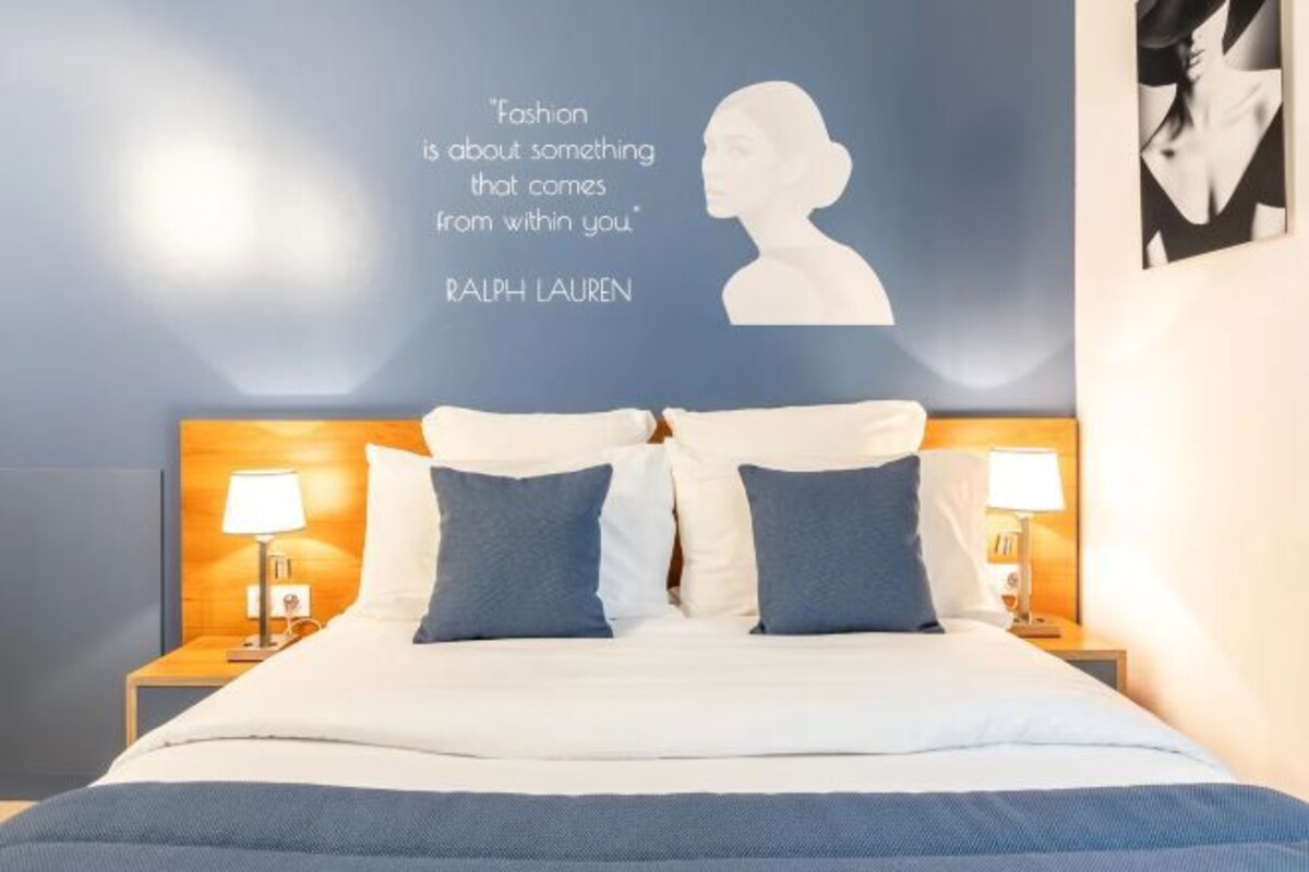 Habitación de hotel con una cama, dos lámparas de noche, una cita de Ralph Lauren en la pared y parte de un cartel.