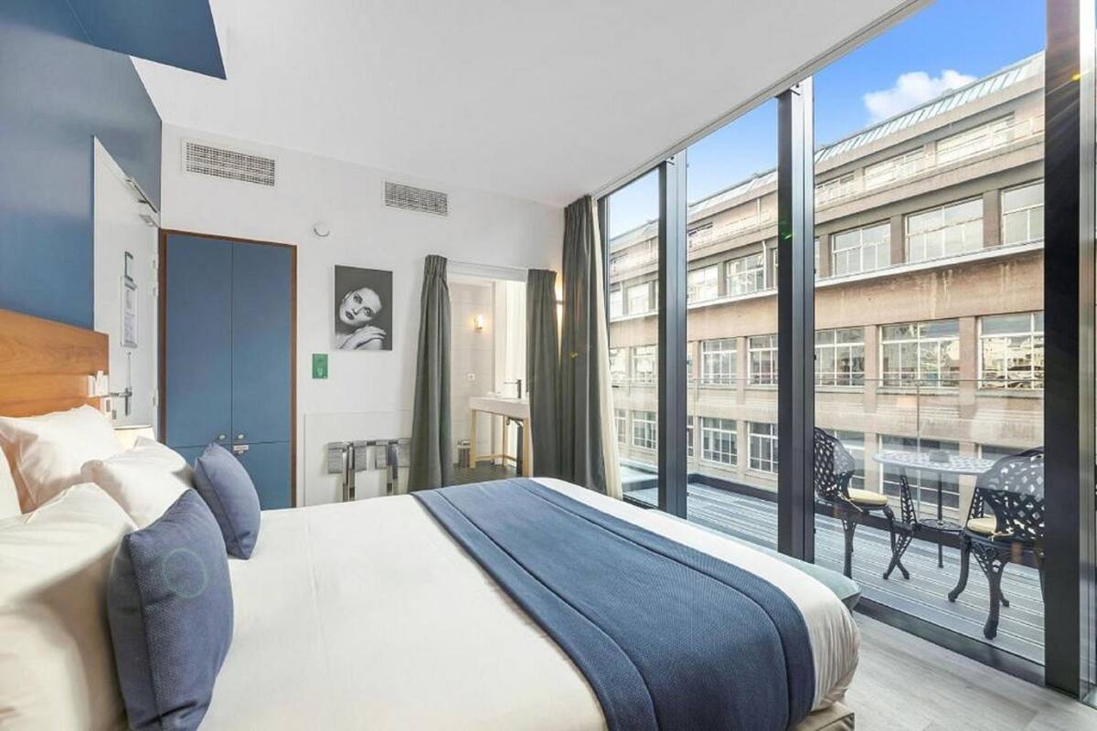 A modern hotel room with a large bed, a desk, and a balcony with outdoor seating.