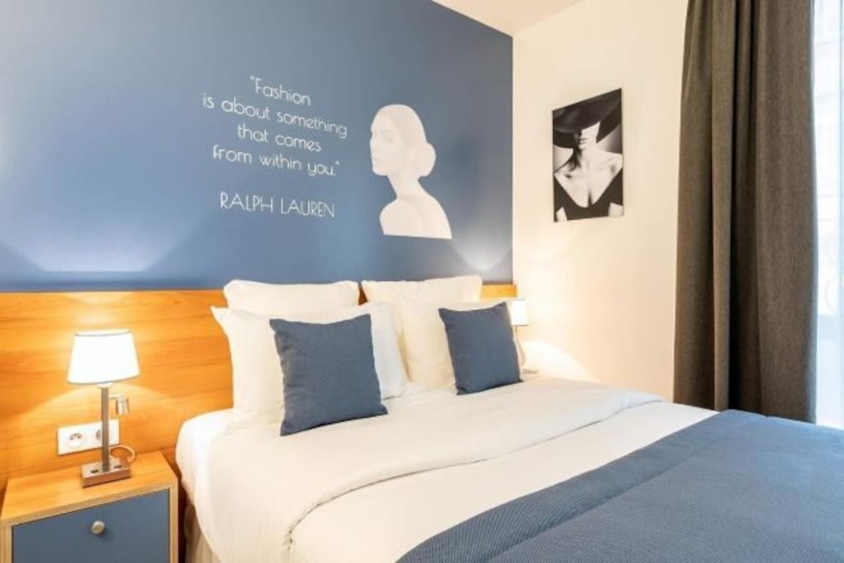 A hotel room with a bed, a nightstand with a lamp, a blue wall with a quote, and a window with curtains.