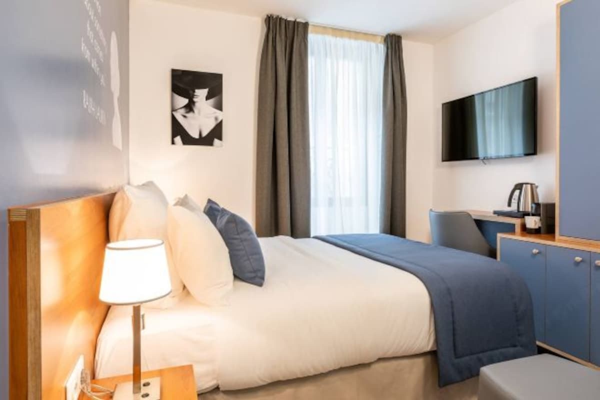 A hotel room with a bed, a desk, a chair, a TV, and a window with curtains.