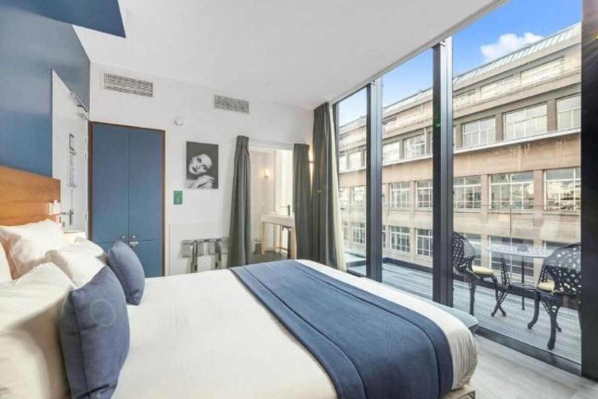 Una habitación de hotel moderna con una cama grande, vistas a la ciudad y un balcón con mobiliario de exterior.