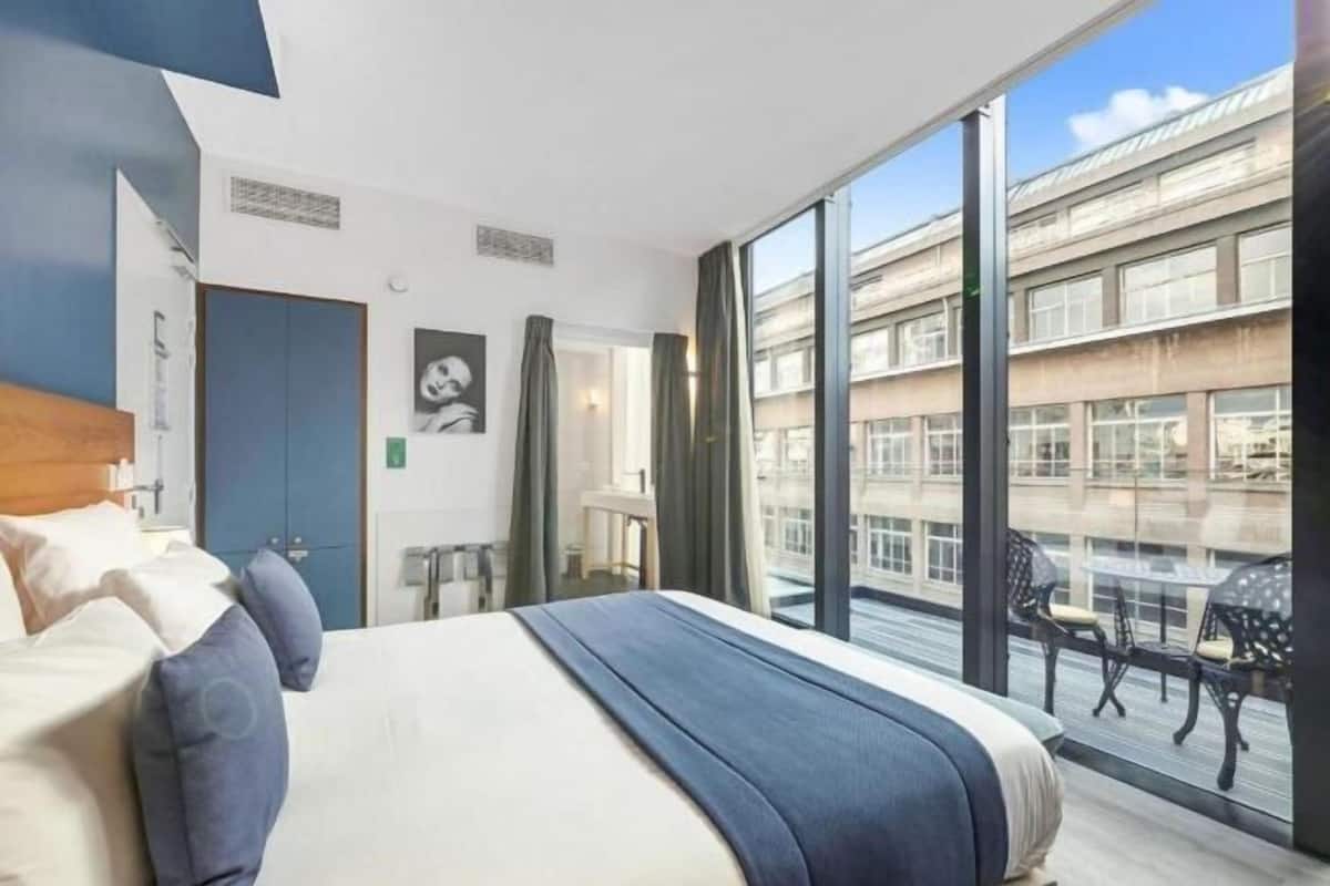 A modern hotel room with a large bed, a view of the city, and a balcony with outdoor seating.