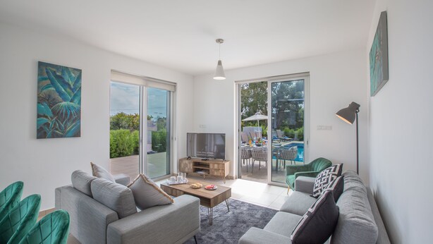 Una sala de estar moderna con un sofá gris, una mesa de centro y un televisor. Posee un ventanal corredero que da a un área exterior con piscina.