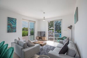 Una sala de estar moderna con un sofá gris, una mesa de centro y un televisor. Posee un ventanal corredero que da a un área exterior con piscina.