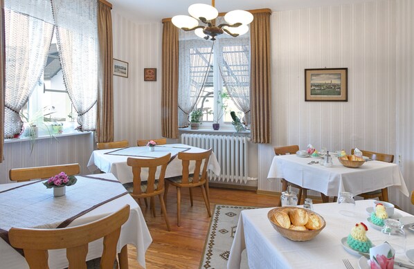 A dining area with wooden tables and chairs, a chandelier, and a view of the city through lace curtains. - Pension Haus am Walde (Brodenbach)