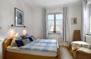 A bedroom with a double bed, two bedside lamps, a window with curtains, a small table with a tablecloth, and a chair. - Pension Haus am Walde (Brodenbach)