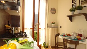 A kitchen with a stove, pots, and fresh vegetables. There is a dining area with a table set for two, a chair, and a clock on the wall.