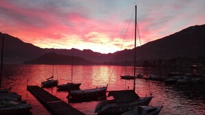 A sunset over a calm lake with boats docked at a pier, surrounded by mountains.