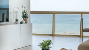 Ein modernes Wohnzimmer mit großem Fenster mit Meerblick, einem Flachbildfernseher, einem runden Holztisch und einem weißen Schrank.