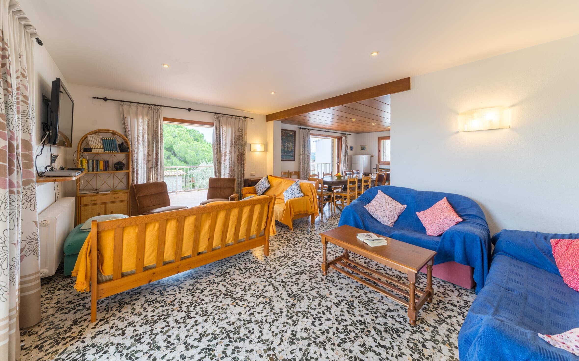 Een gezellige woonkamer met een houten bank, een blauwe bank en een houten tafel. Er is een balkon met uitzicht op groen.