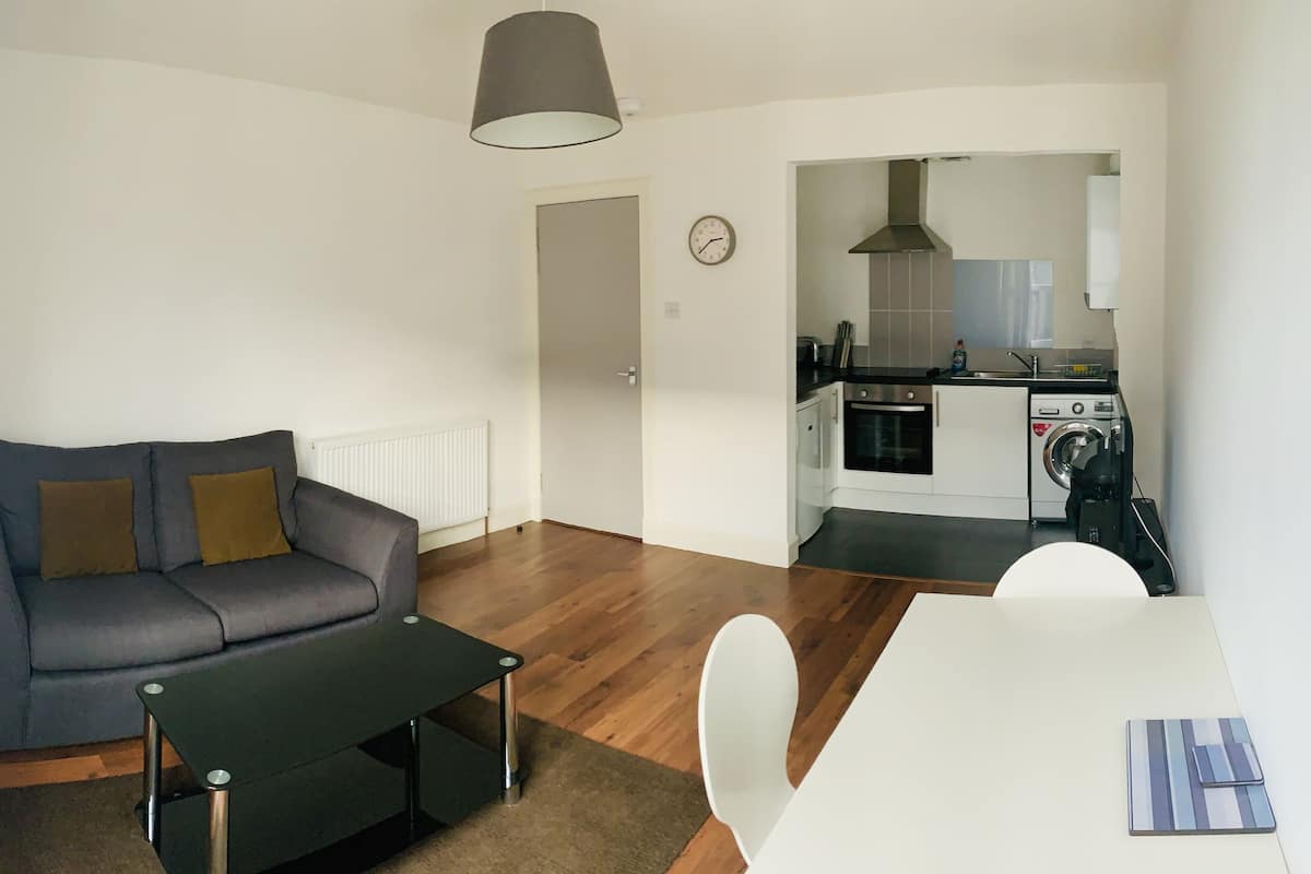 Ein modernes Wohnzimmer mit einem grauen Sofa, einem schwarzen Couchtisch und einem weißen Esstisch. Im Hintergrund ist eine Küche mit Herd, Backofen und Waschmaschine zu sehen.
